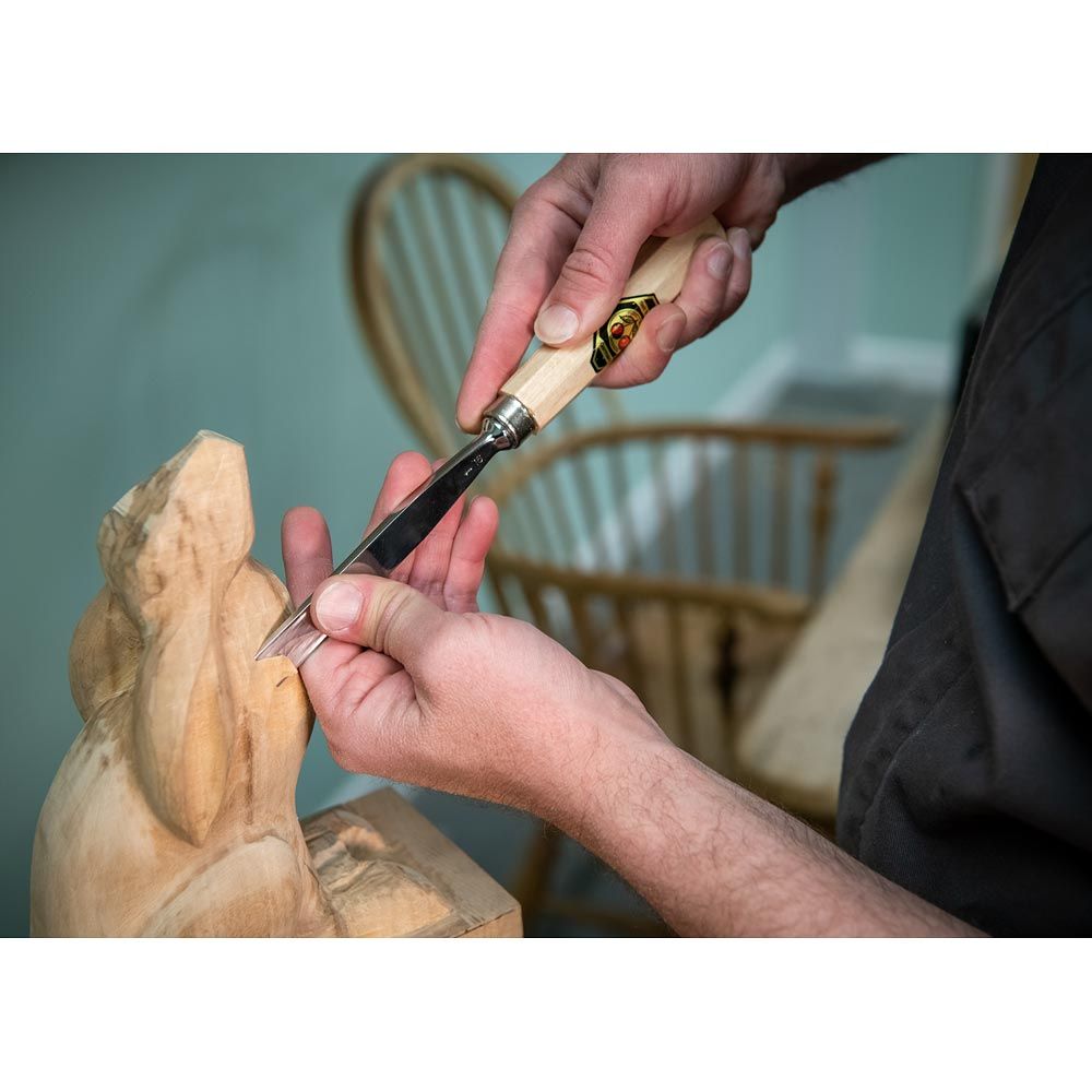 Person carving a wooden sculpture with a chisel, blurred chair in the background