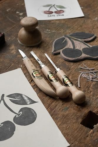Three carving tools on a wooden surface with carved cherry designs and a paper with cherries.