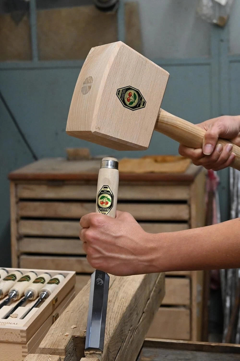 Person holding a wooden mallet with a visible brand logo about to strike the back of a chisel in a workshop setting.