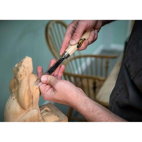 Person carving a wooden sculpture with a chisel, blurred chair in the background